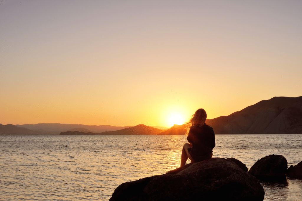 Woman watching over the sea at sunset to represent letting go of limiting stories so you can transform your career
