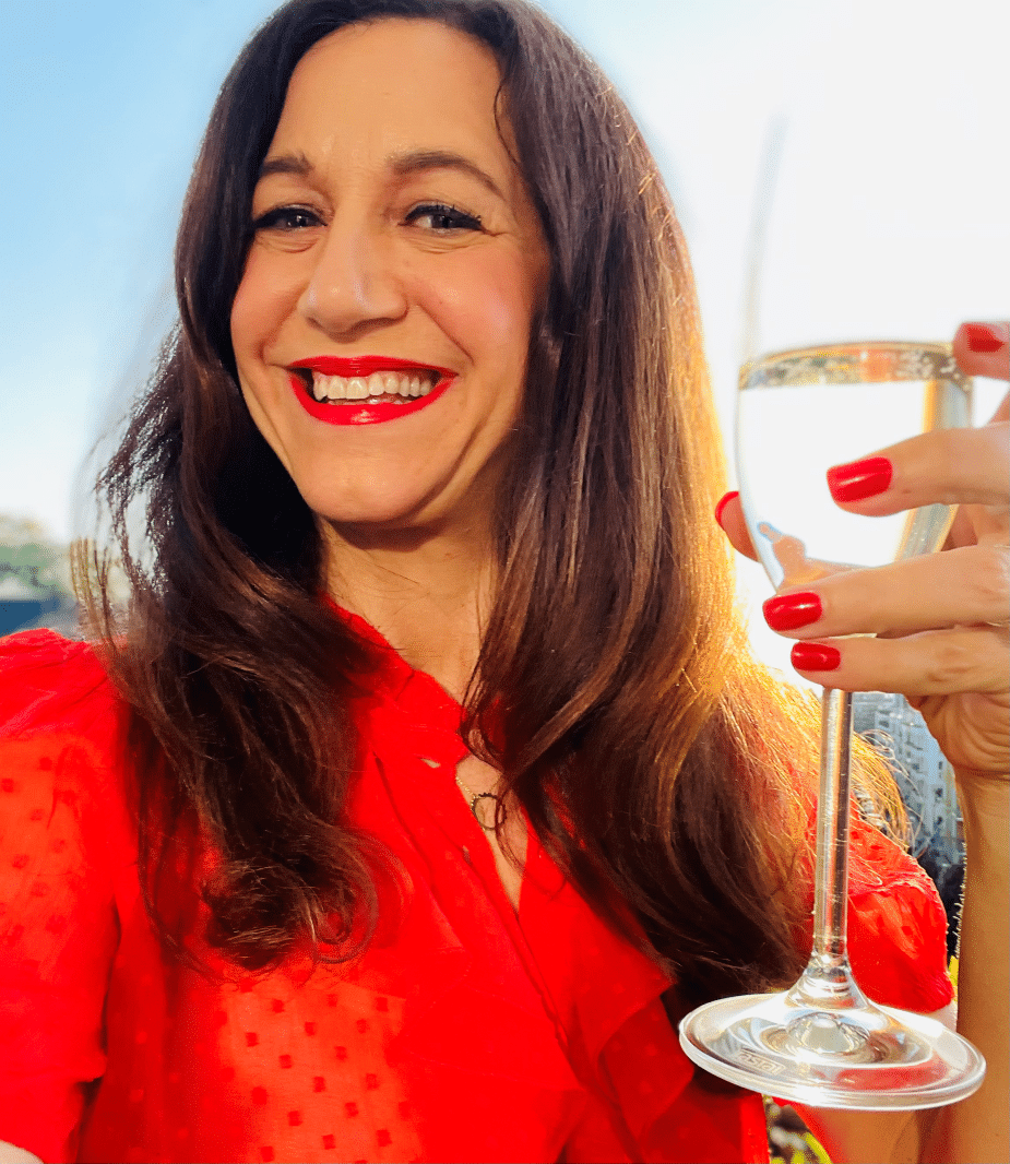 Life coach Katarina Stoltz in a red blouse holding champagne as she smiles in celebration at the camera