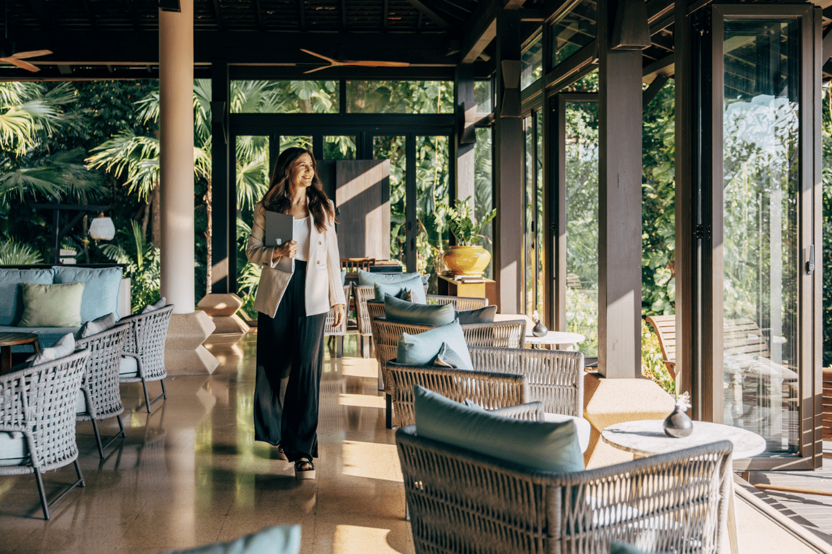 Life coach Katarina Stoltz walks through a bright, open-air lounge with wicker chairs and large windows, holding a tablet, with lush greenery visible outside.