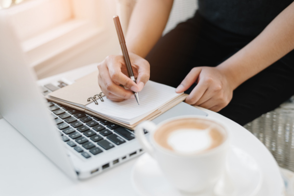 Woman writing yearly goals in a diary with an open laptop and a cup of coffee.