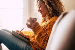 Woman in yellow jumping sitting and drinking coffee whilst journalling.